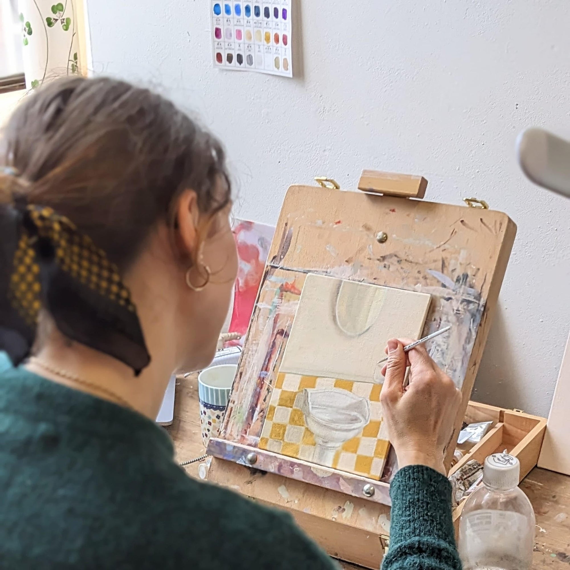 A woman sitting and painting a sink on her easel, with a dark yellow and beige/white checkerboard background and a mirror.