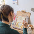 A woman sitting and painting a sink on her easel, with a dark yellow and beige/white checkerboard background and a mirror.